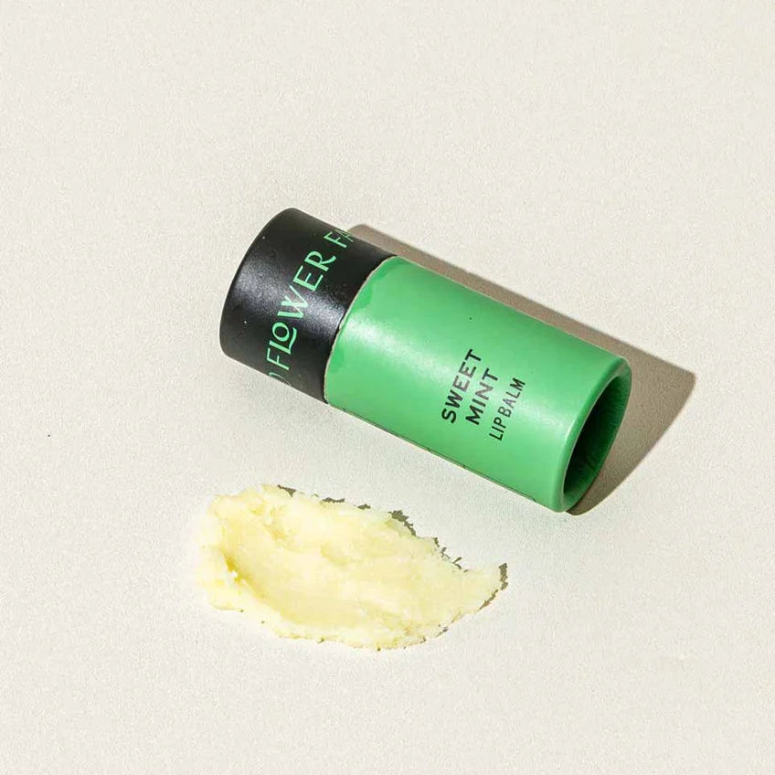 A green and black tube of Flower 1 Sweet Mint Lip Balm lies on its side next to a smear of pale minty cream on a light beige background.