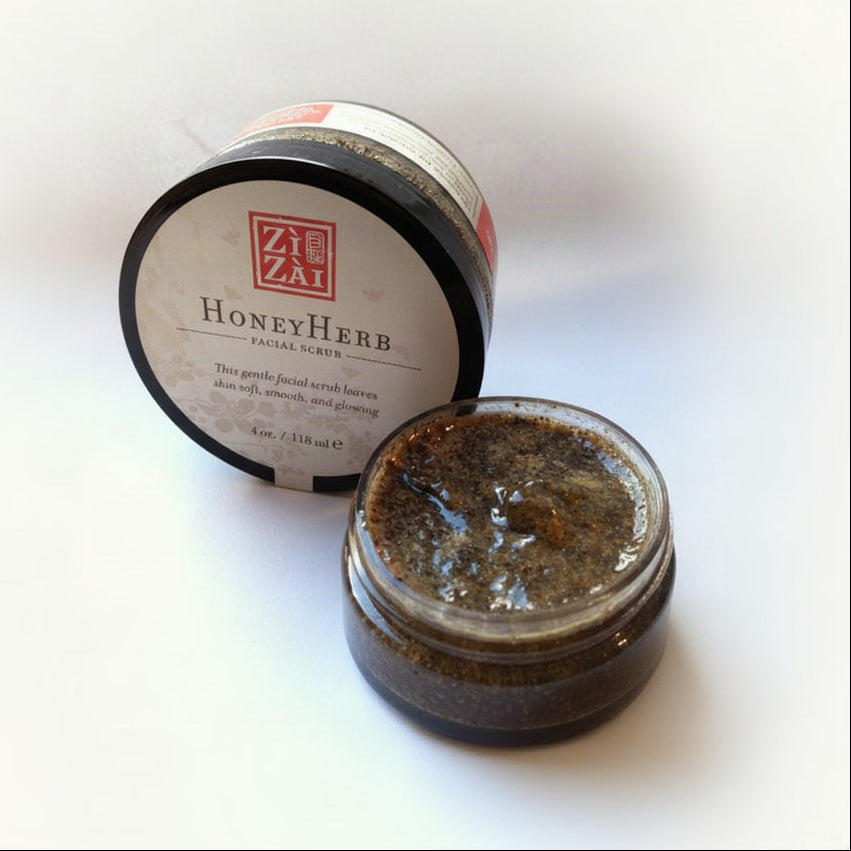 A jar of Honey Herb Facial Scrub with its lid open, revealing a dark, grainy product. The label on the lid includes the brand 'Zi Zai Dermatology' and the product name 'Honey Herb Facial Scrub' with additional text describing the product's benefits.