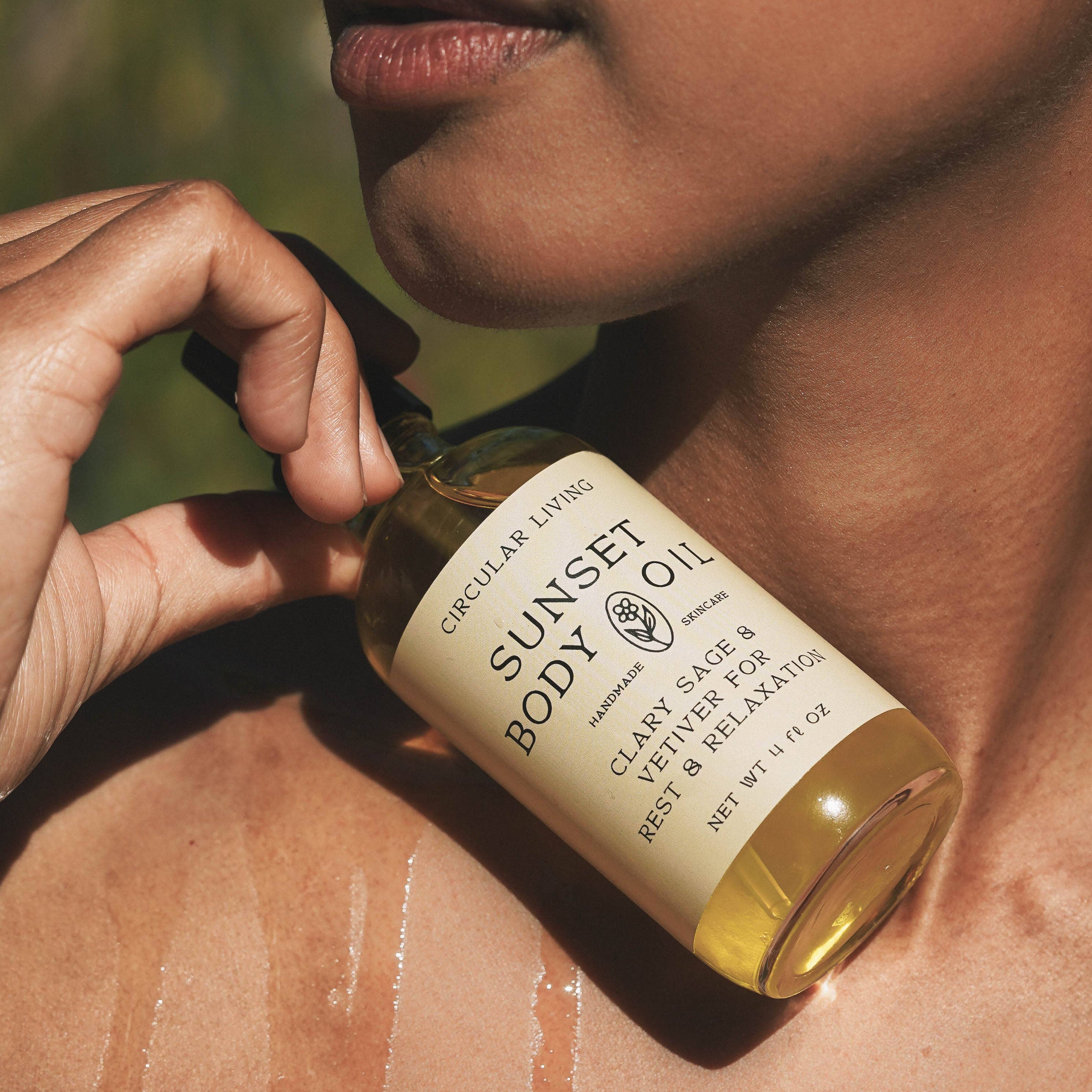 Person holding a bottle of 'Sunset Body Oil' against a natural background