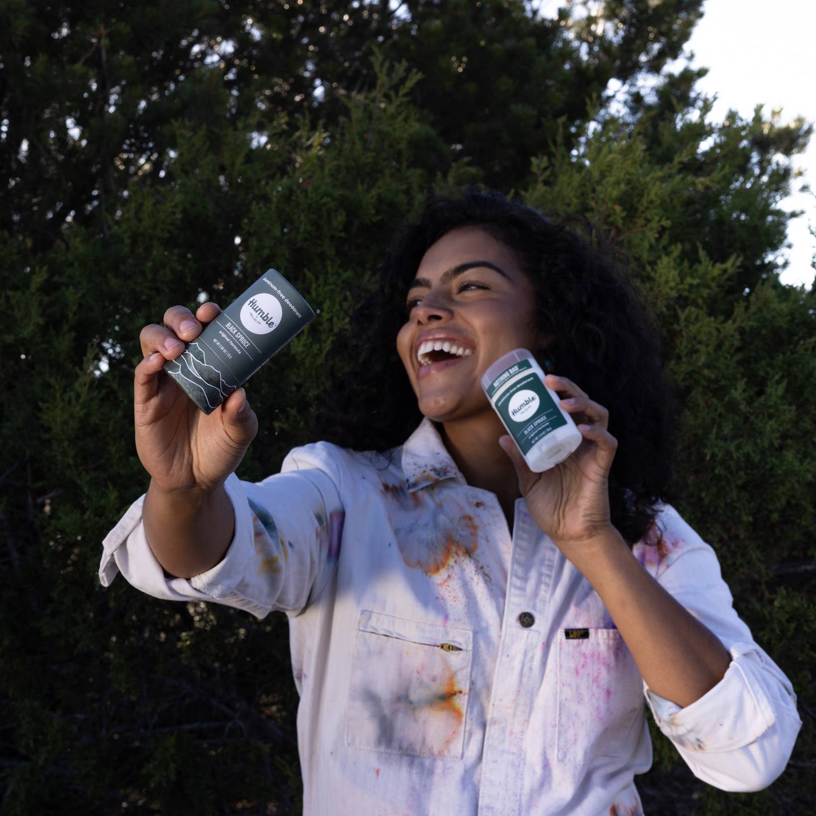 Woman holding two skincare products outdoors with greenery in the background