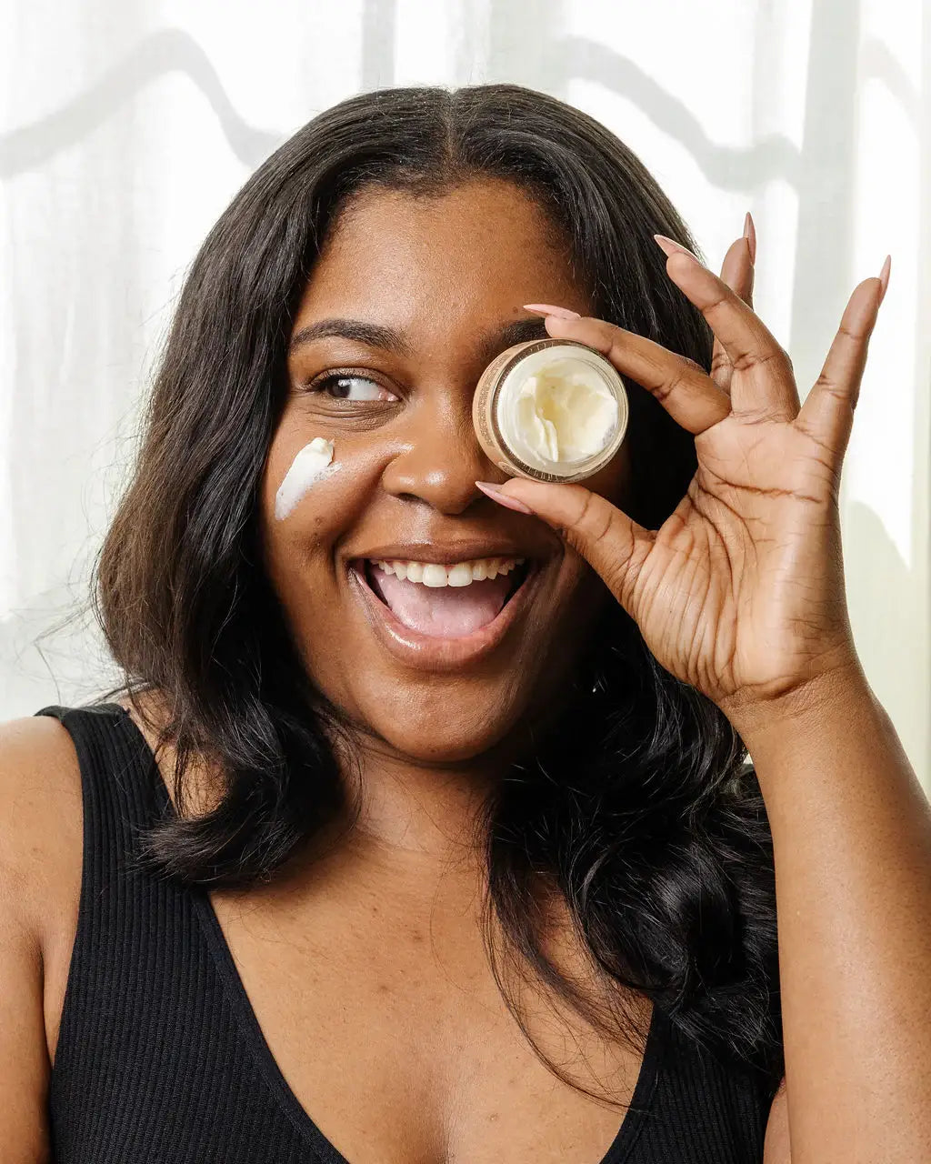 Woman holding a jar of cream near her eye with a smile, against a neutral background