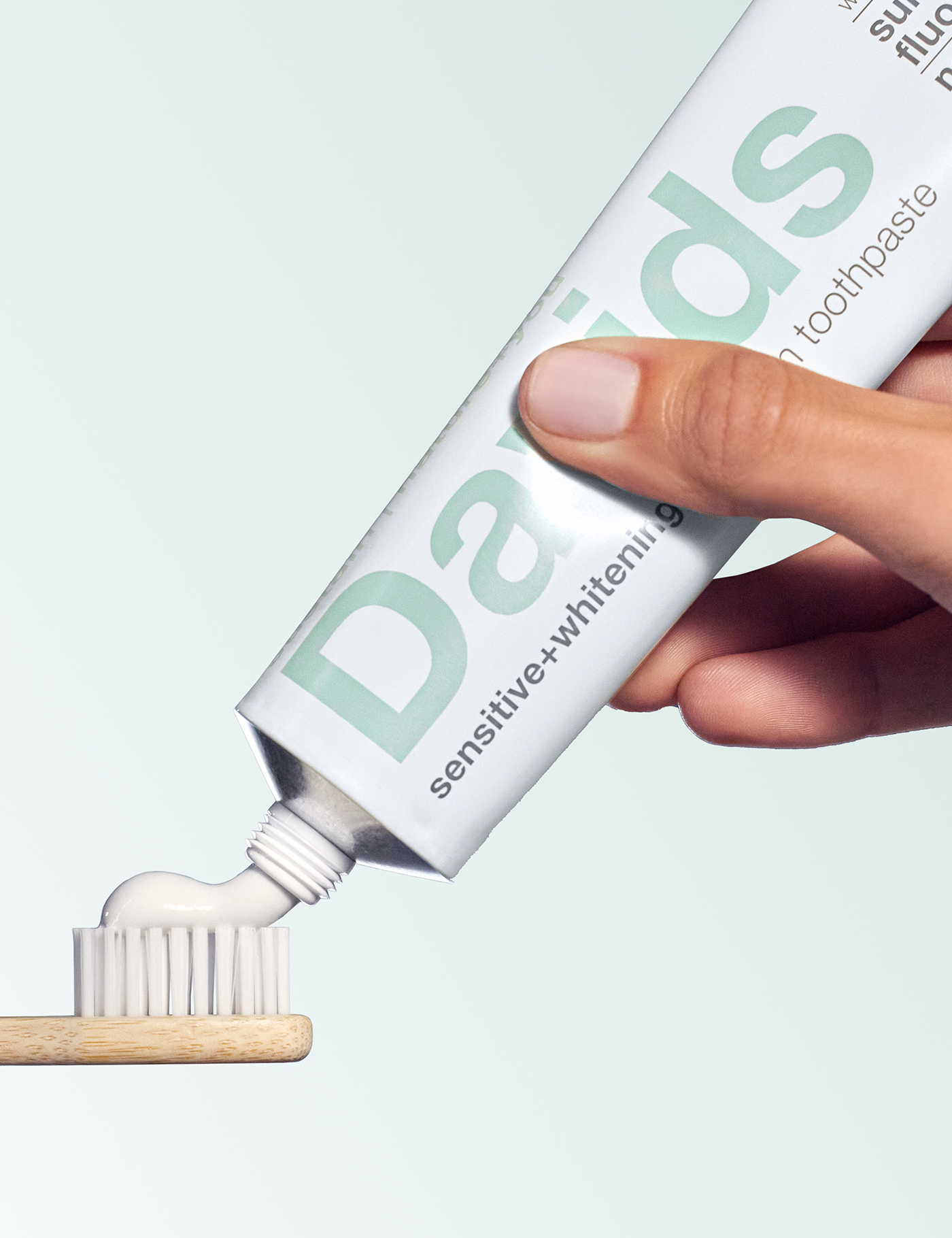 Hand applying Dabudub toothpaste to a wooden toothbrush against a light green background
