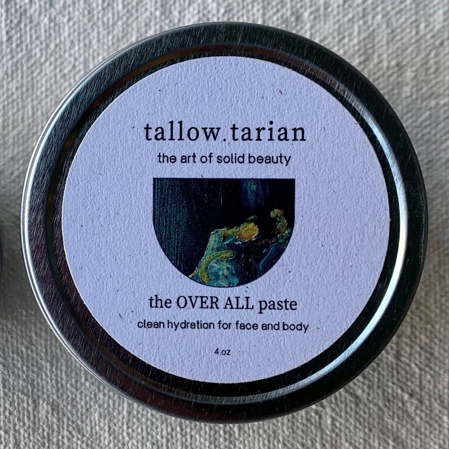 Two jars of Tallowtarian Over All Paste with labels showing the product name and an illustration of a face, one jar with a metallic lid and the other with a fabric lid.