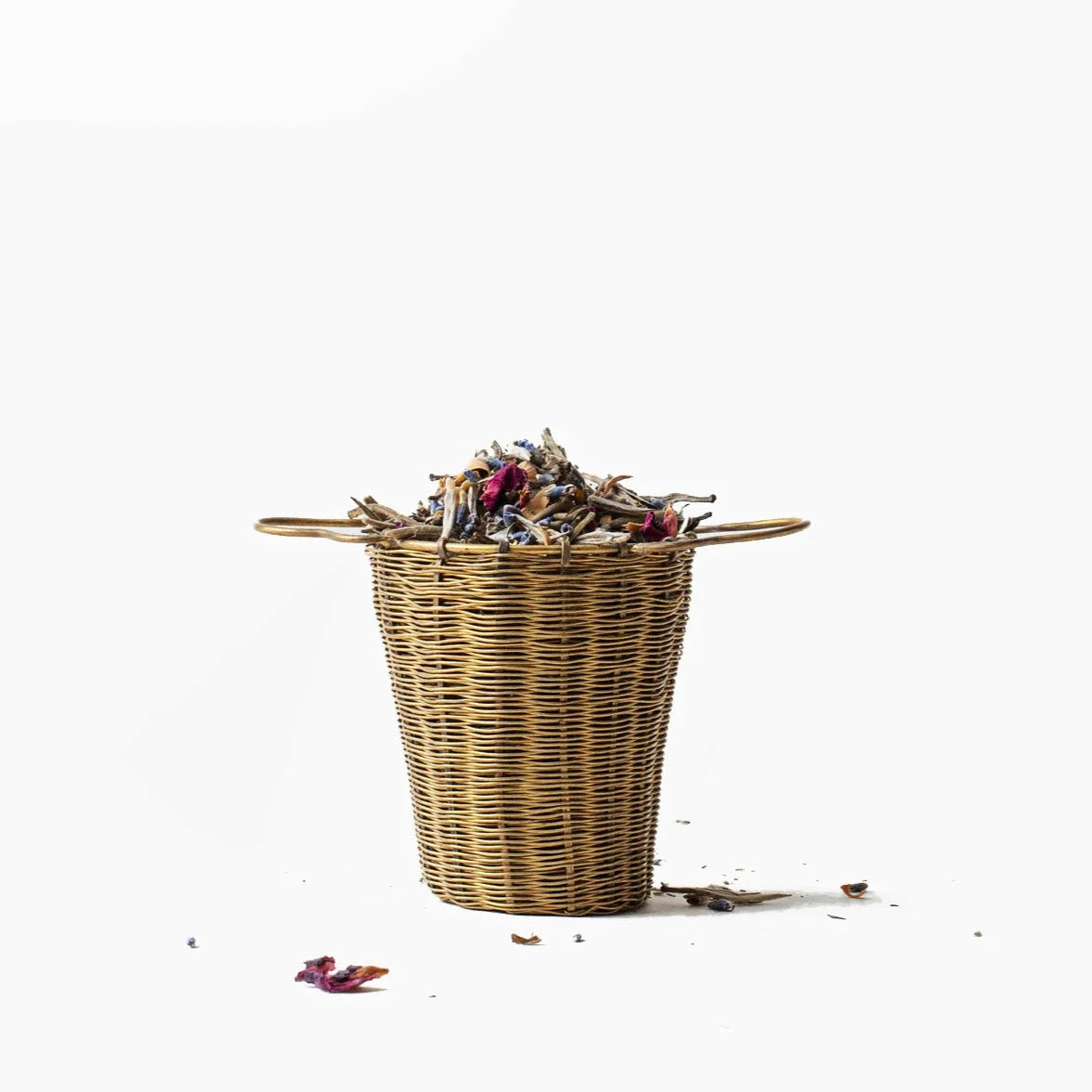 Brass woven tea strainer holding dried herbs and leaves on a white background