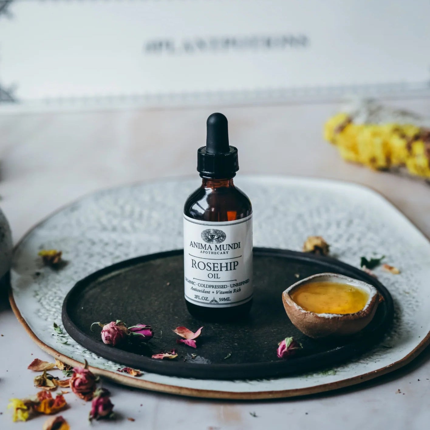 Bottle of Anima Mundi Rosehip Oil on a black tray with dried flowers and a small dish on a marble surface.