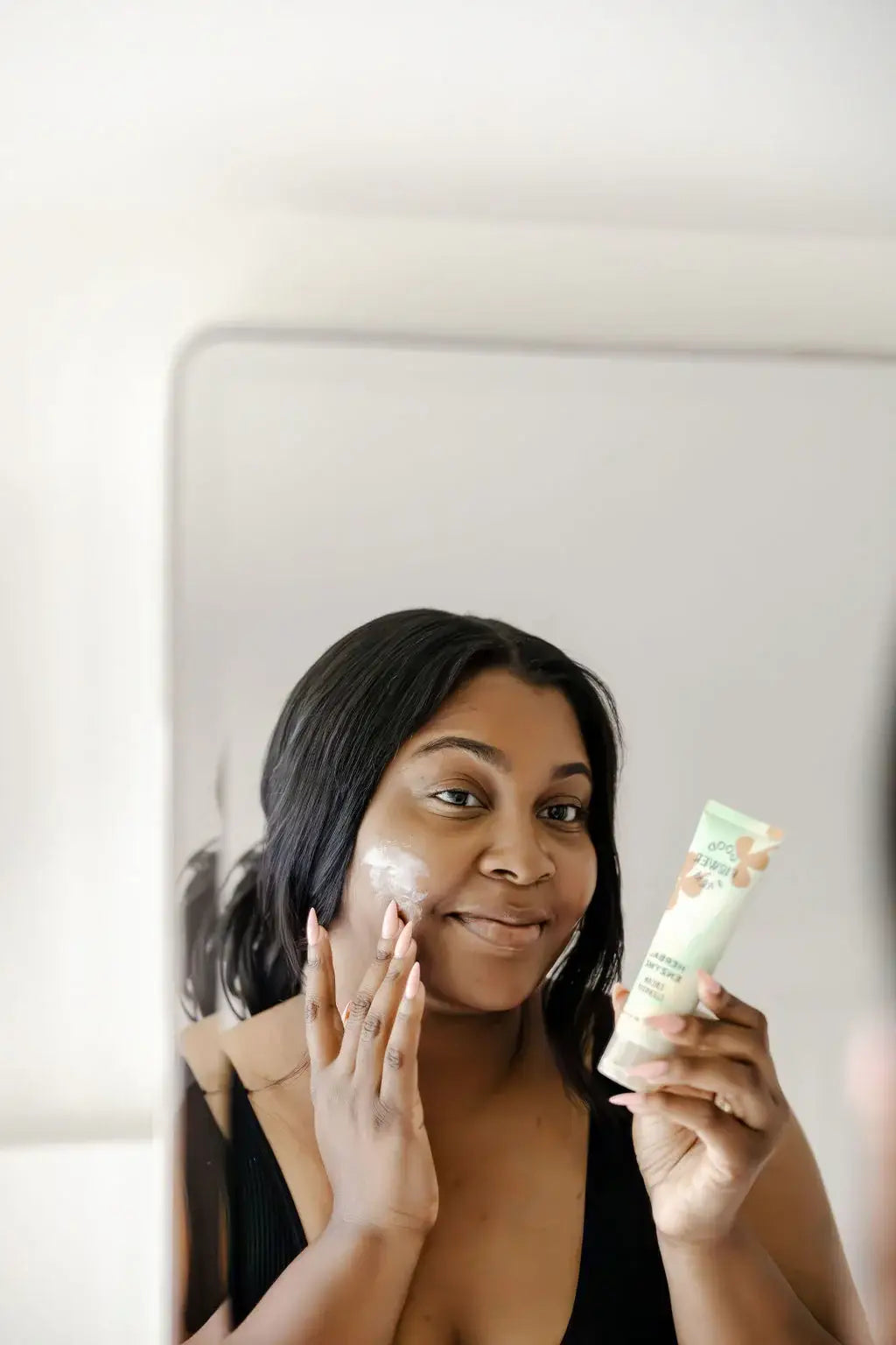 Woman applying cream to her face with a tube of cream in front of a mirror.