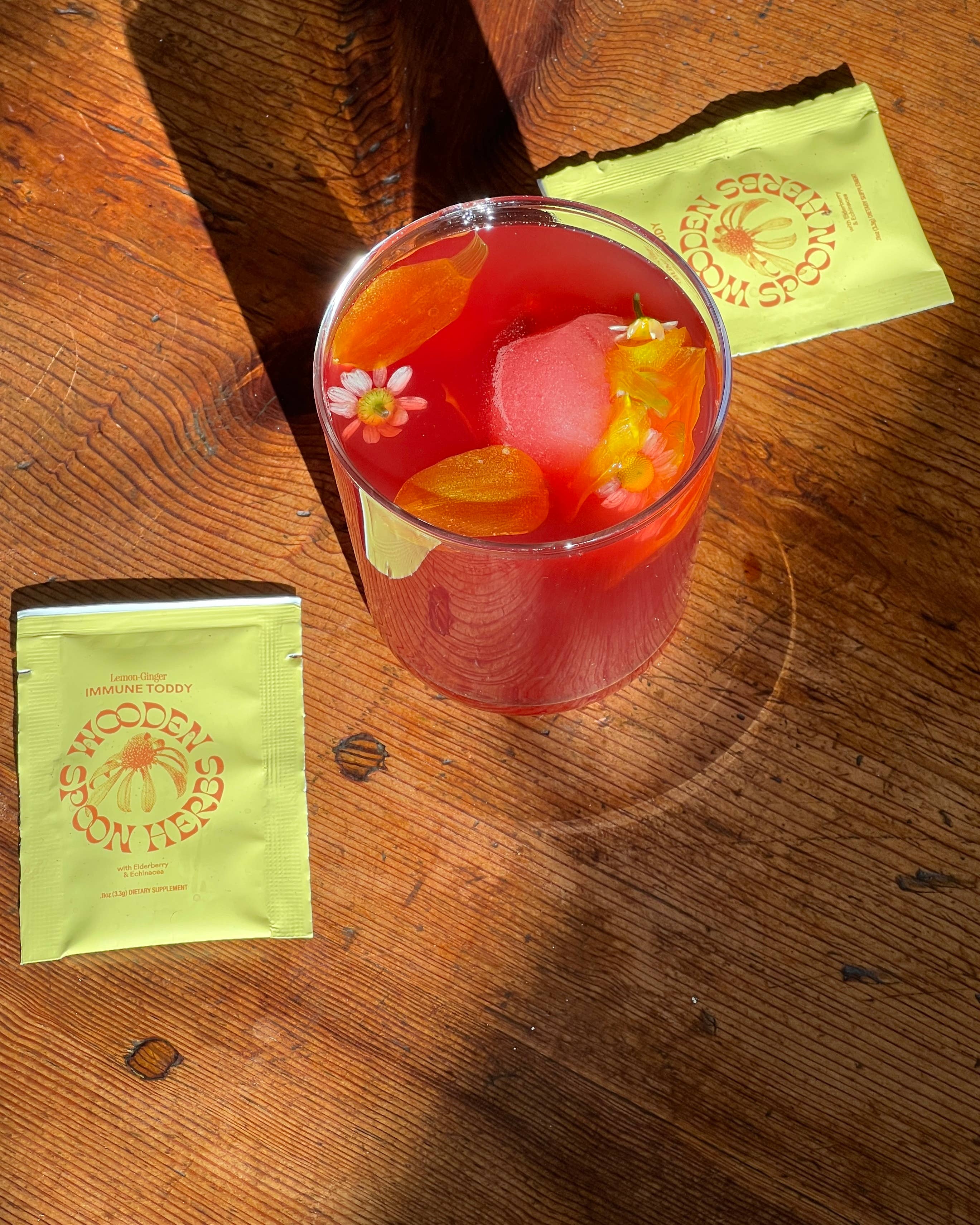 Glass of red iced tea with floral elements on a wooden surface, accompanied by two green packets.